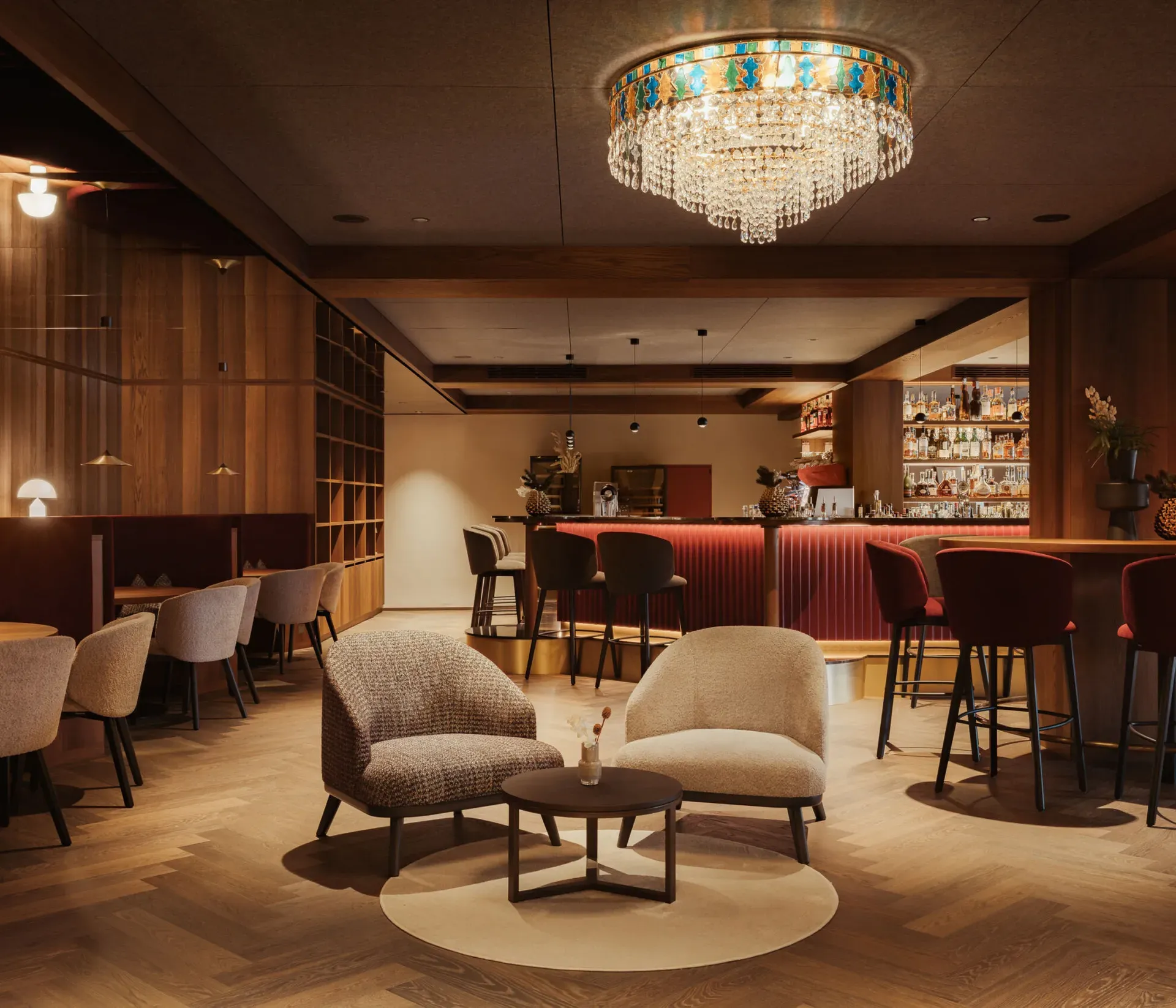 Warmly lit hotel bar with plush seating, a round table, and a chandelier. A wooden bar with red chairs creates a cozy, inviting atmosphere.