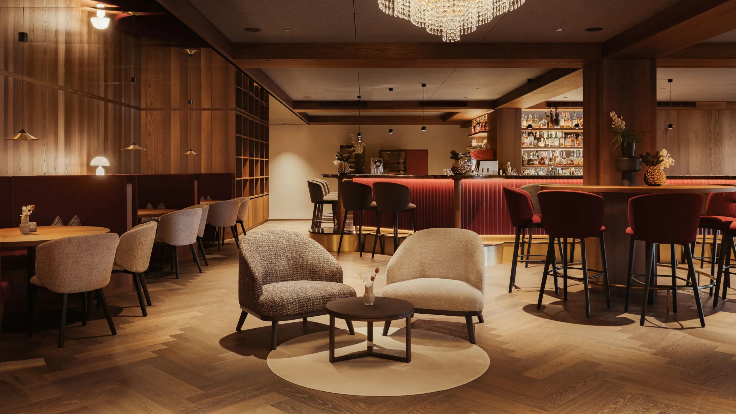 Warmly lit hotel bar with plush seating, a round table, and a chandelier. A wooden bar with red chairs creates a cozy, inviting atmosphere.