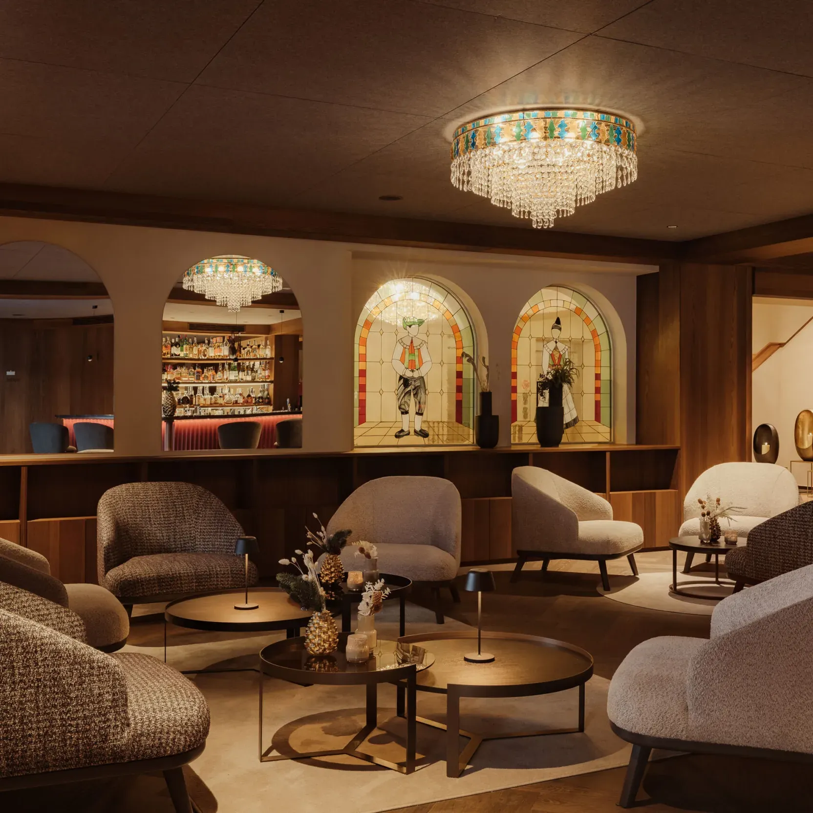 Cozy lounge with soft gray chairs around round tables, under a sparkling chandelier. Arched mirrors and a lit bar create a warm, inviting atmosphere.
