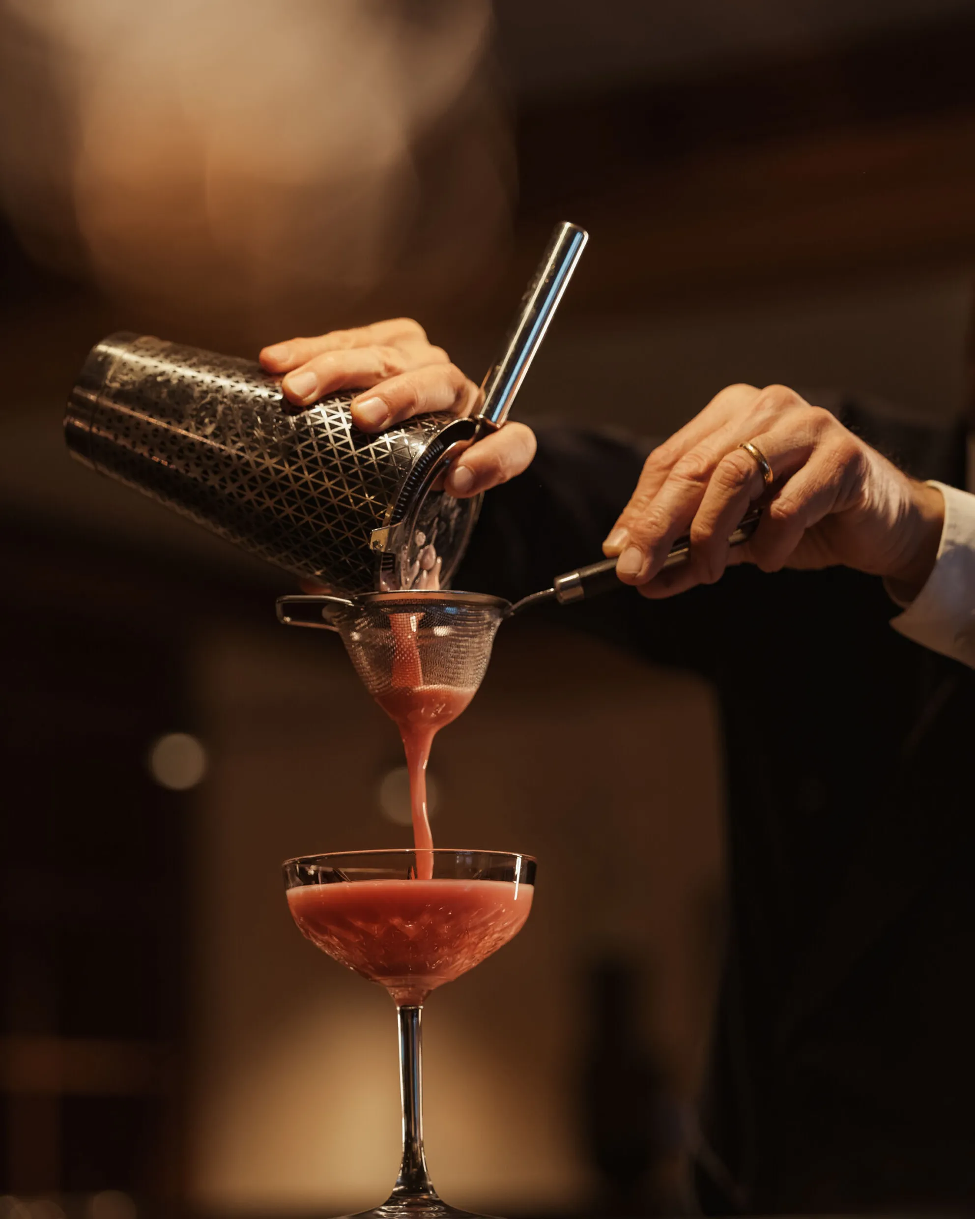 Ein Barkeeper gießt einen leuchtend pinken Cocktail in ein Glas und verwendet dabei Shaker und Sieb. Die warme Beleuchtung erzeugt eine elegante, anspruchsvolle Atmosphäre.