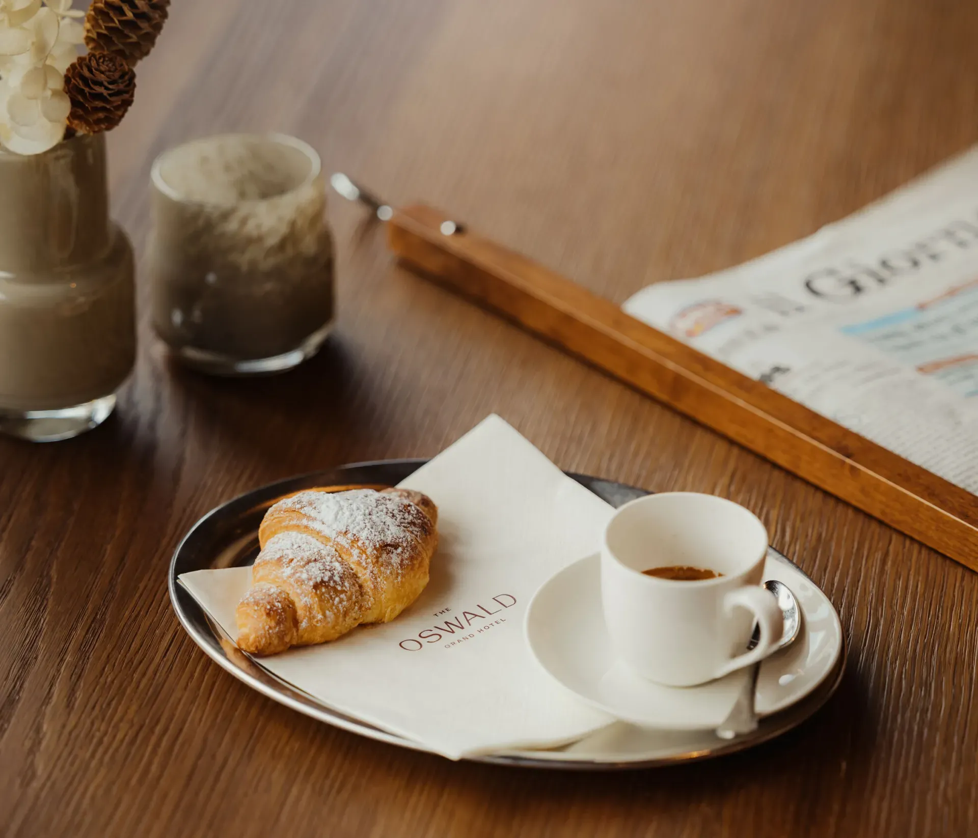 Ein Croissant auf einer Serviette mit einer Espressotasse liegt auf einem Tablett auf einem Holztisch. Eine Zeitung und dekorative Vasen in der Nähe schaffen eine gemütliche Atmosphäre.