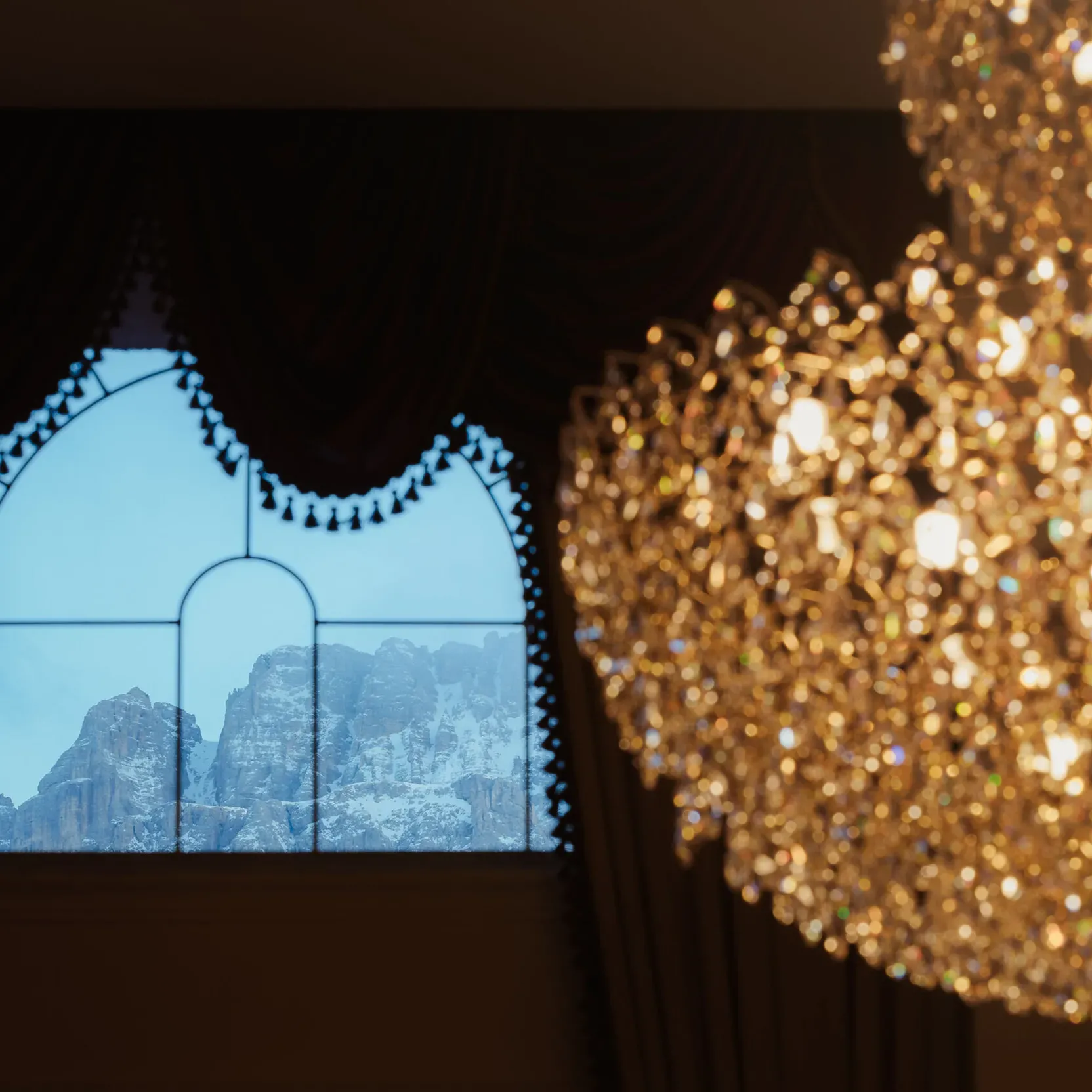 A large crystal chandelier dominates the right side of the image, while a window with an ornate curtain reveals a snowy mountain view, creating a contrast between elegance and nature.