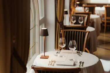 Elegant restaurant interior with a round table set for two, featuring wine glasses, cutlery, and a small table lamp. The atmosphere is cozy and intimate.