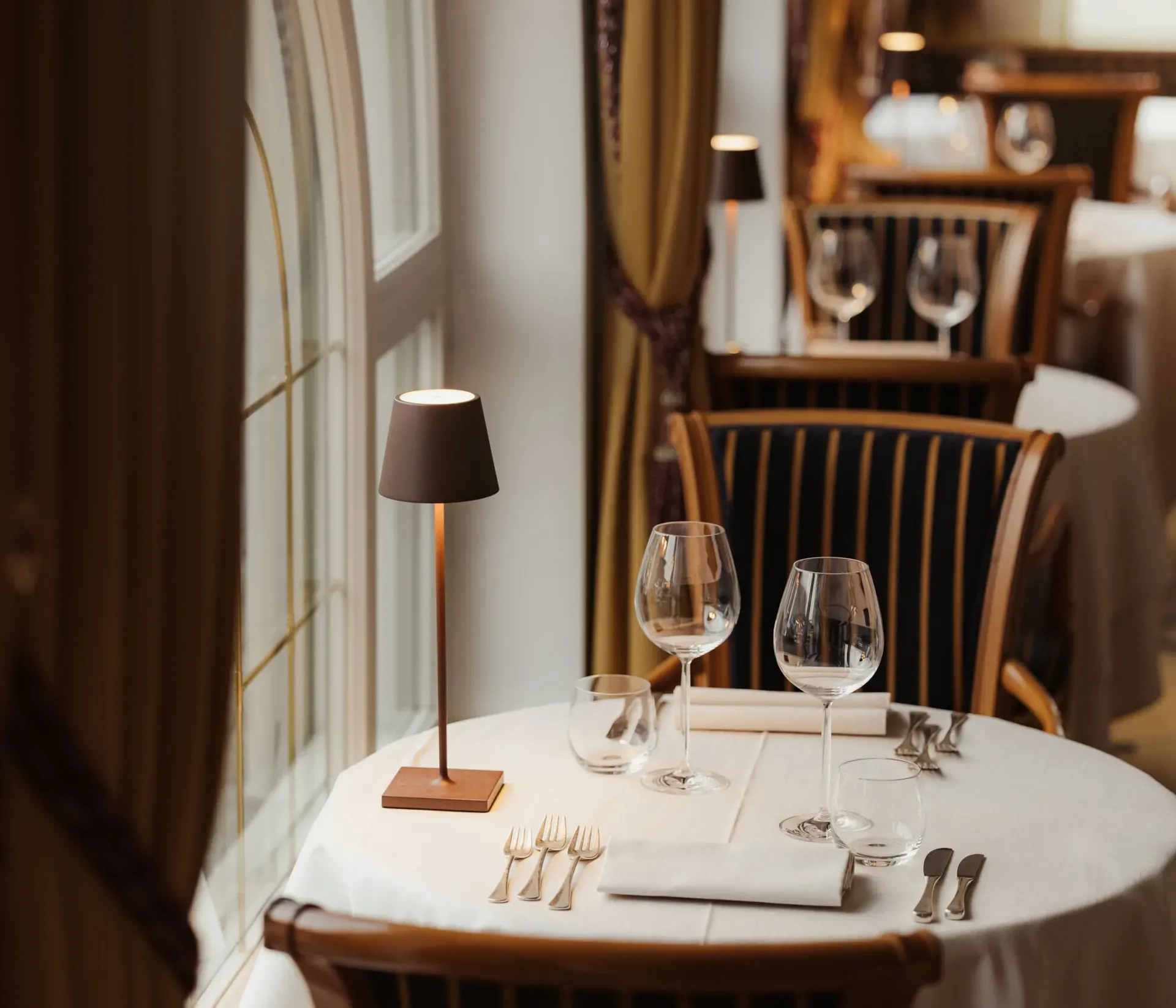 Elegantes Restaurantinterieur mit einem runden Tisch für zwei Personen, ausgestattet mit Weingläsern, Besteck und einer kleinen Tischlampe. Die Atmosphäre ist gemütlich und intim.