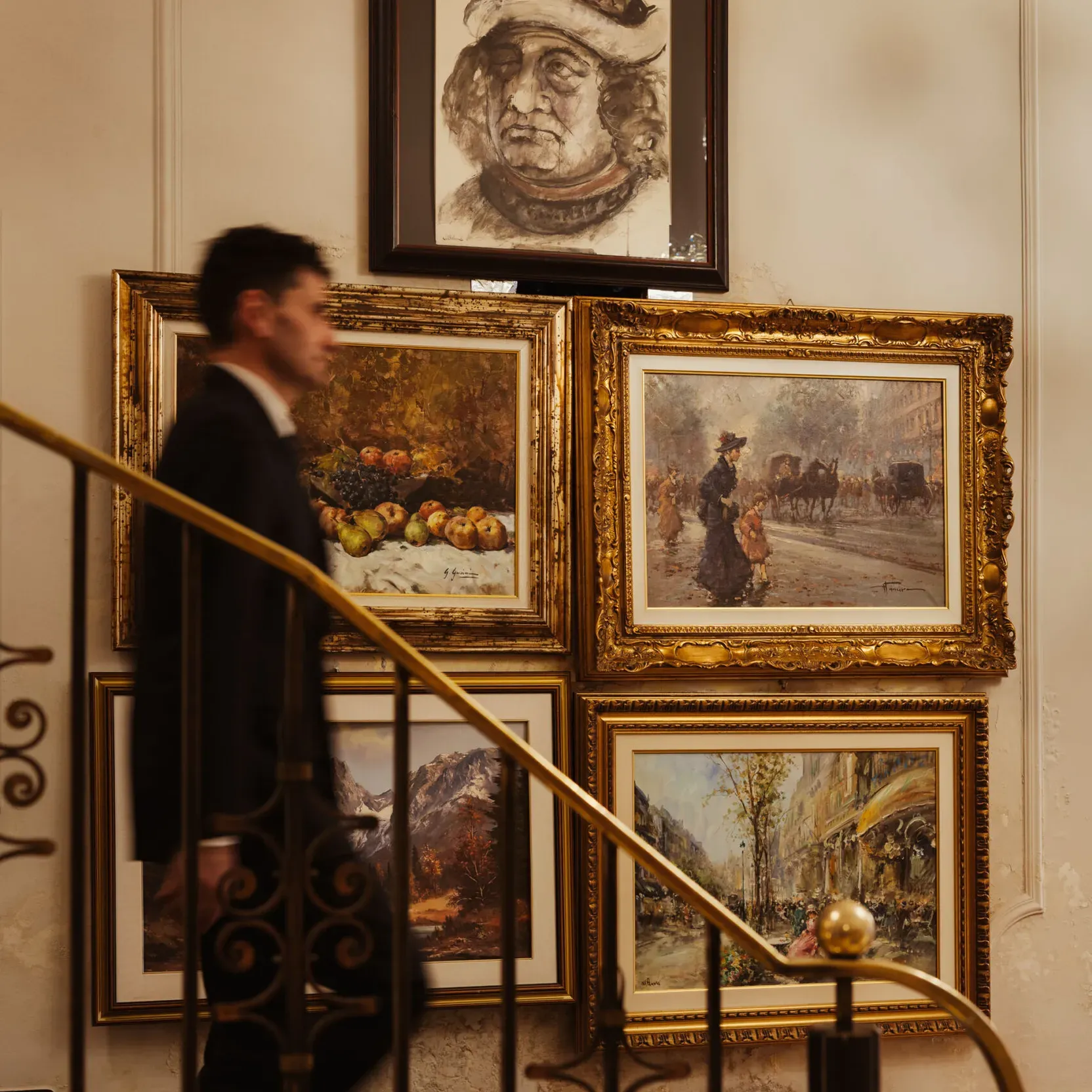 A man in a suit ascends a curved staircase adorned with ornate railings. Behind him, framed paintings depict various scenes, adding an elegant, artistic ambiance.