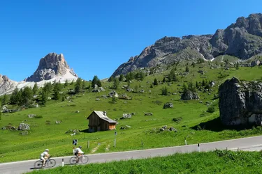 Two cyclists climbing a mountain pass in the Dolomites, with a green meadow, a hayloft, and a snow-capped mountain in the background.