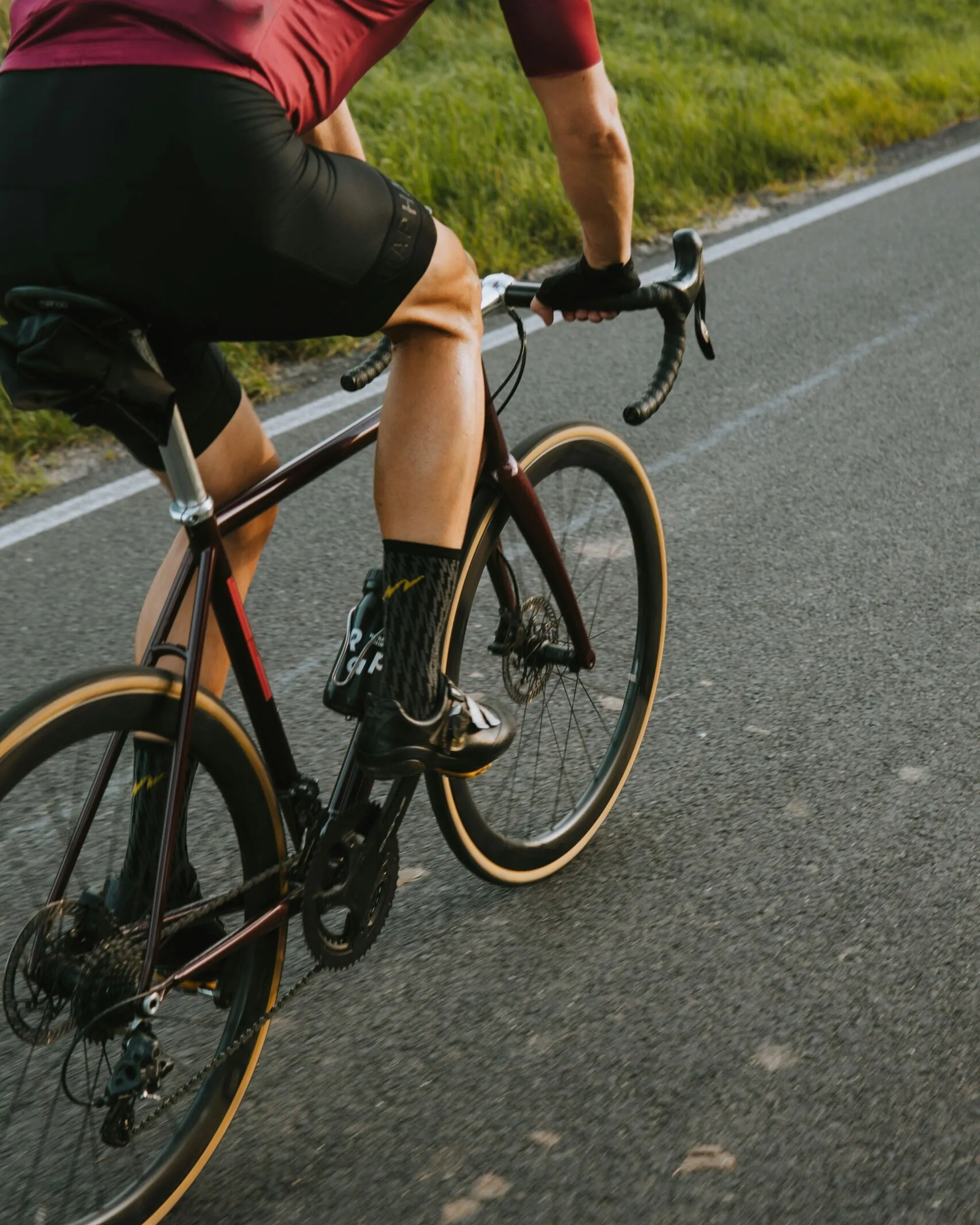 Cropped cyclist pedaling along a mountain road, wearing a burgundy jersey and black shorts, with a grassy slope at the roadside.