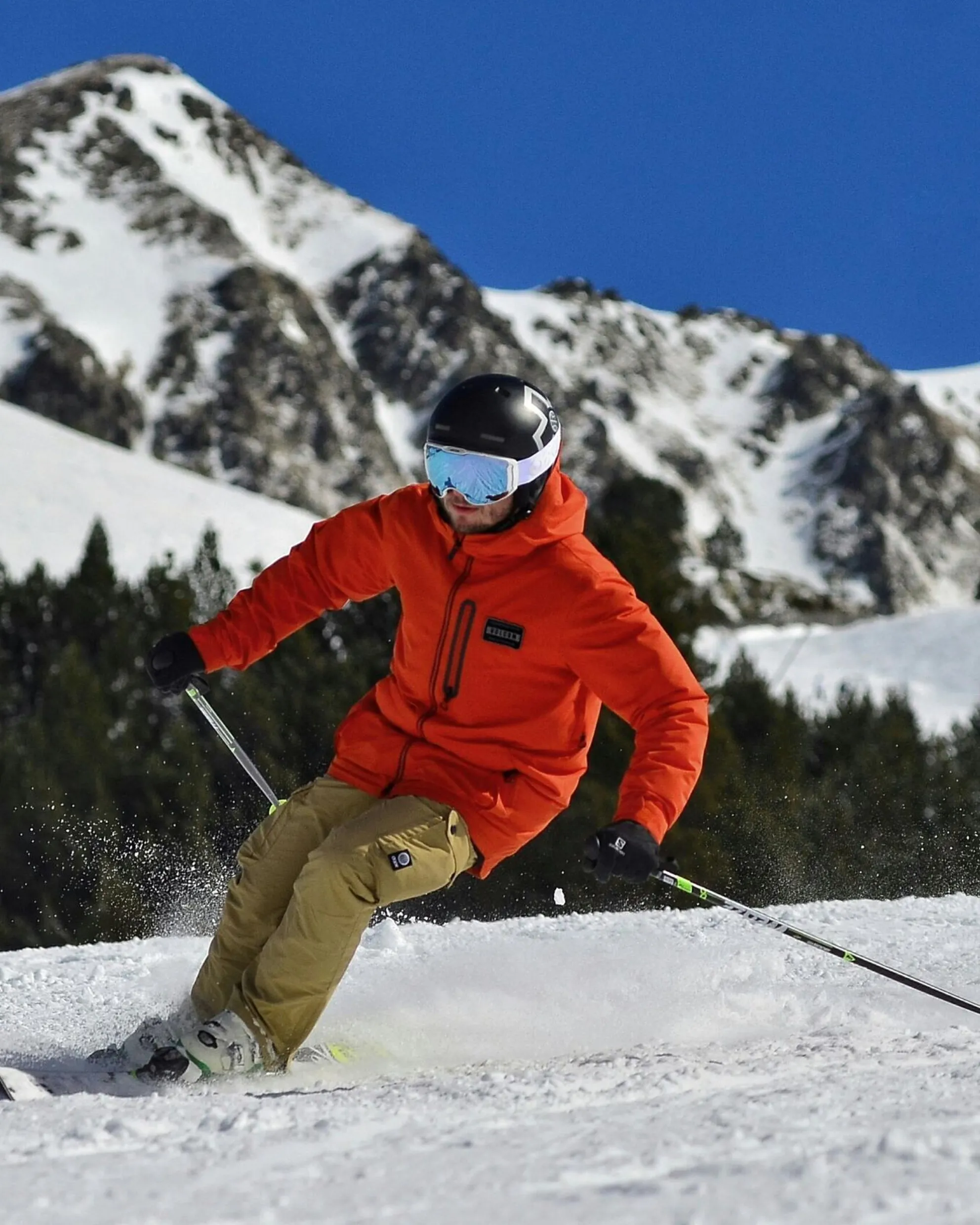 Sciatore che conduce una curva sugli sci, indossando giacca arancione, pantaloni beige, casco e maschera, con sullo sfondo montagna innevata e bosco durante una giornata di sole invernale.