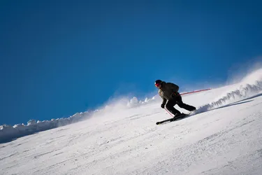 Sciatore con tuta verde militare che conduce una curva sulla pista da sci, sollevando leggermente la neve durante la discesa.
