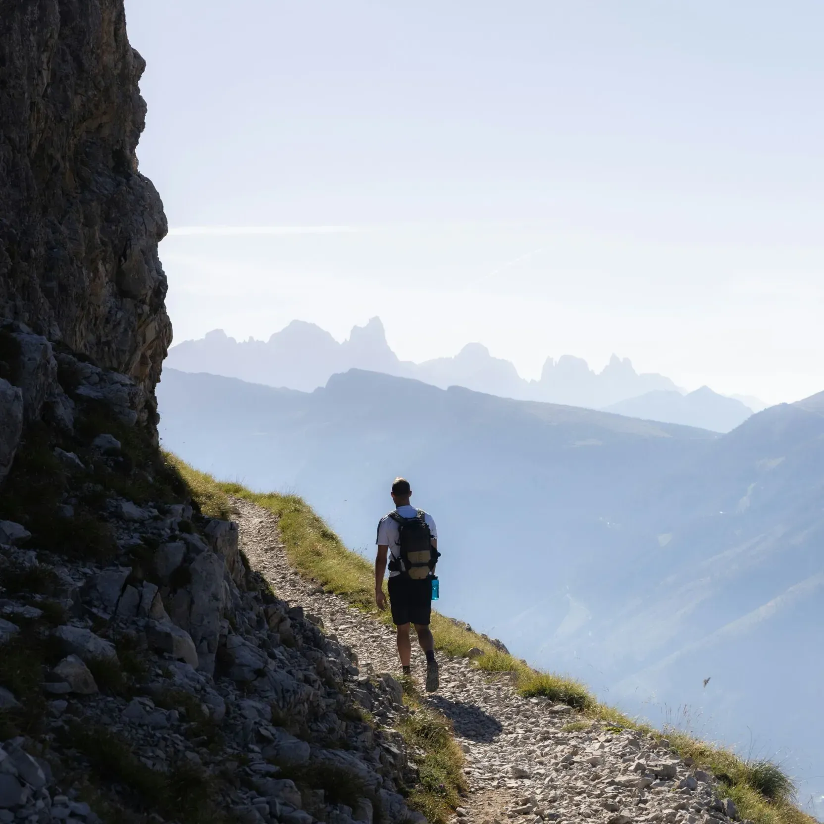 Man walking along a mountain trail with a rocky wall on the right casting a shadow on the path, backlit, with a mountain range in the background.