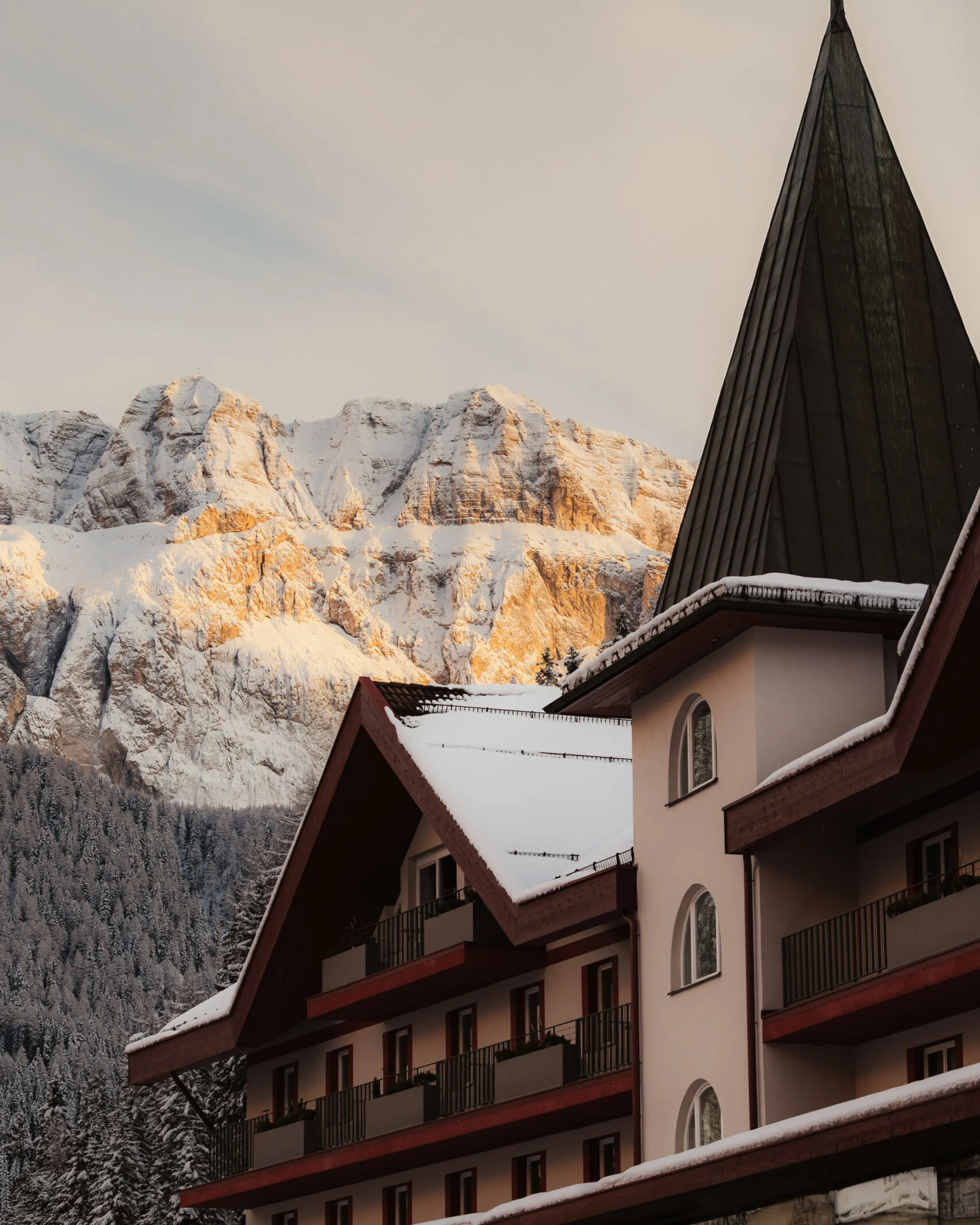 Snow-covered alpine hotel with a steep roof sits in front of sunlit, snowy mountains. A tranquil, wintry atmosphere is captured at sunset.
