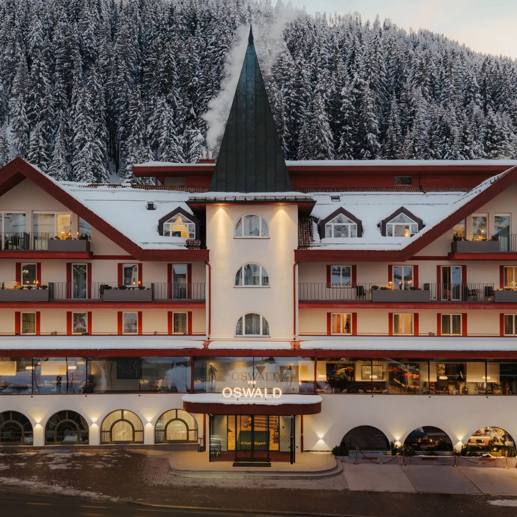 A cozy multi-story hotel with a central tower and snowy roof, set against a backdrop of snow-covered pine trees, exudes a warm, inviting ambiance.