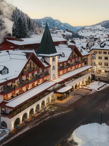 A snowy alpine village at dusk, featuring a grand hotel with pointed roof and arched windows. Warm lights inside create a cozy, inviting atmosphere.