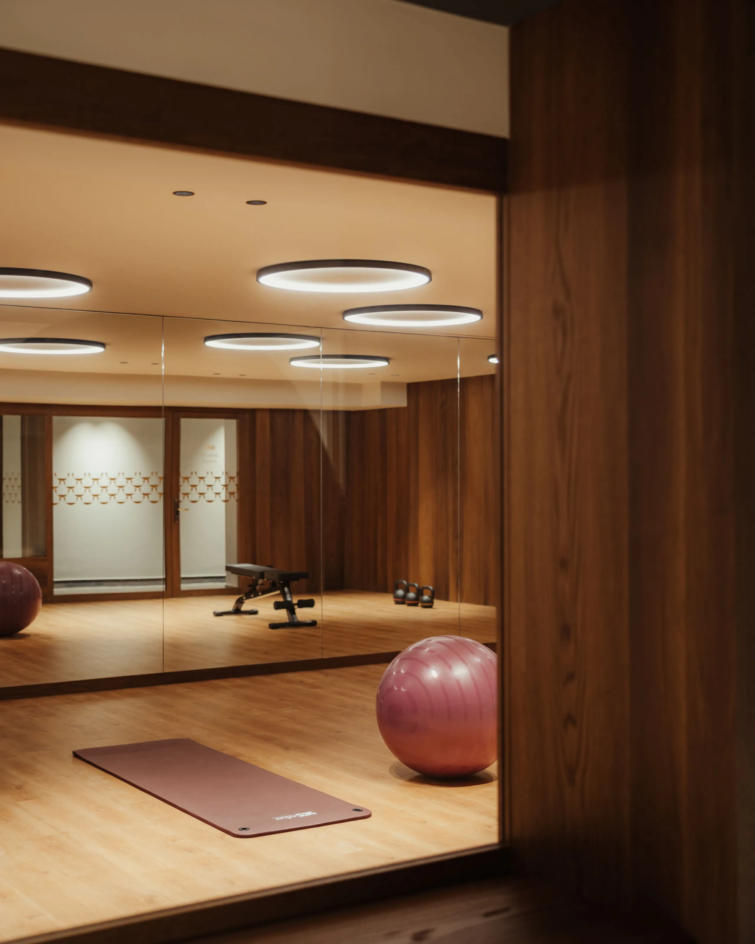 Modern gym with wooden walls and flooring, featuring a yoga mat, exercise ball, and mirrored walls reflecting sleek round ceiling lights.