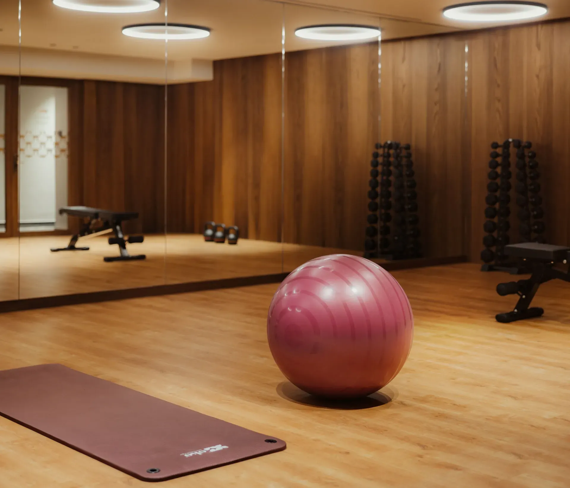 A serene gym with wooden floors, a large purple exercise ball, and a burgundy mat. Dumbbells and benches are reflected in mirrored walls, lit warmly.