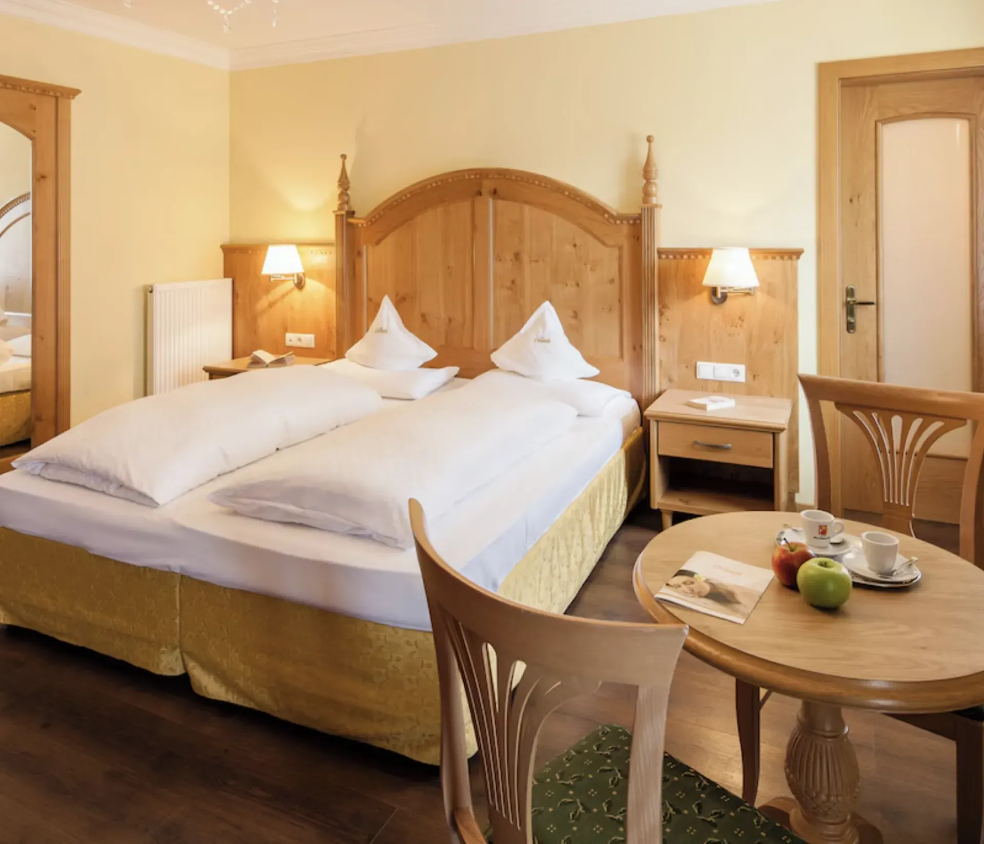 Cozy hotel room with a wooden double bed, crisp white linens, and warm lighting. A round table hosts tea, an apple, and a plate, next to decorative chairs.