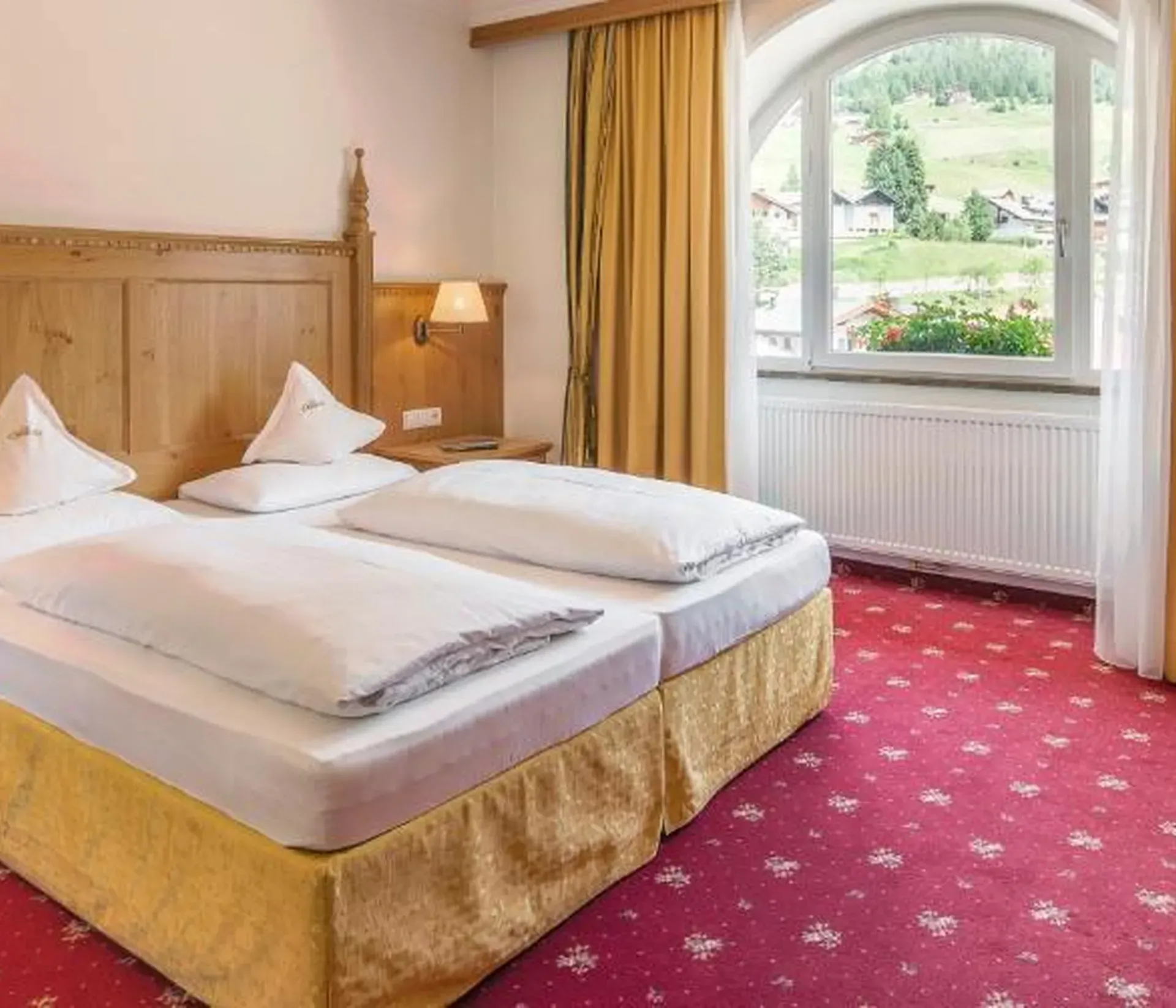 Camera storica dell’Hotel Oswald in legno di cirmolo con moquette rossa, tende drappeggiate in tessuto giallo, luci delle abat-jour accese e vista sul bosco.