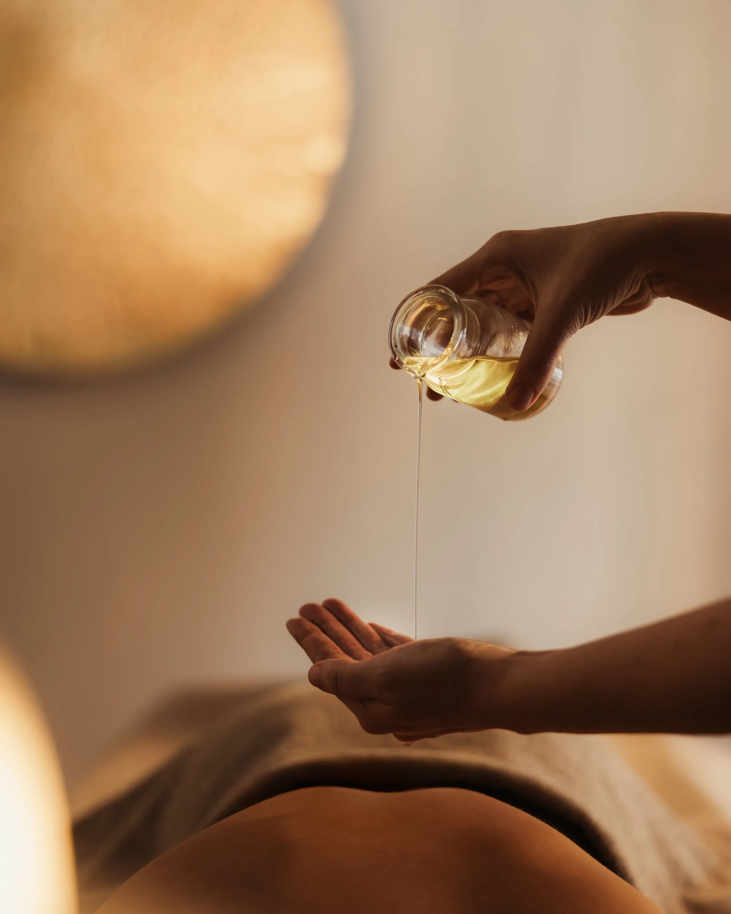 A person's hands pour massage oil onto their palm above a client's back, covered with a towel. Soft lighting creates a warm, relaxing ambiance.