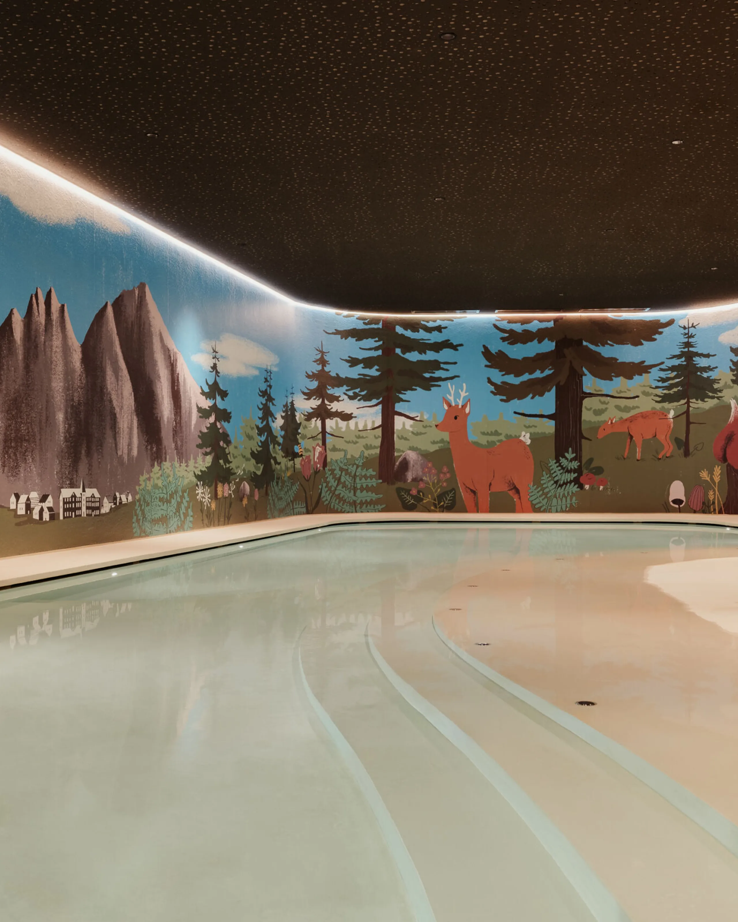 Piscina interna con illuminazione soffusa. Le pareti sono decorate con un murale raffigurante montagne, alberi e animali. Giochi colorati come un dinosauro blu e un canguro rosso circondano la piscina.