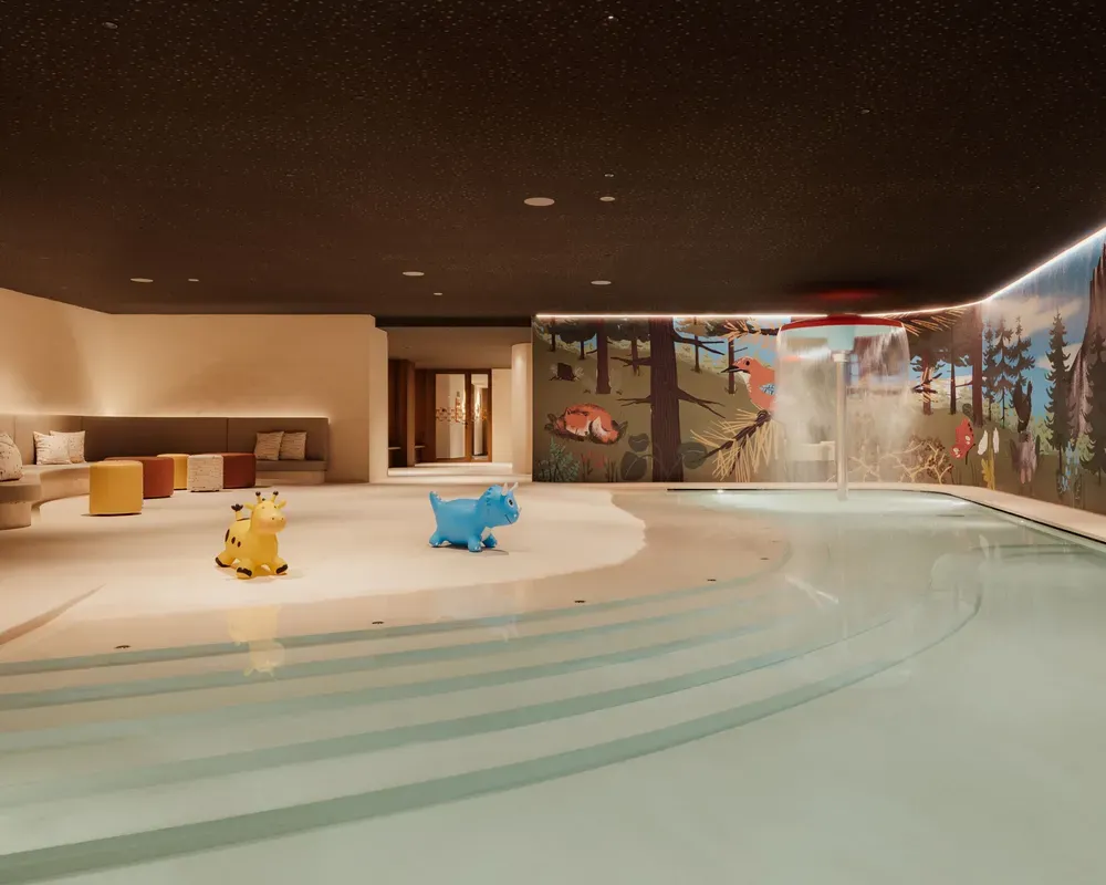 Indoor pool area with playful murals and soft lighting. Features a shallow pool, two colorful animal sculptures, and cushioned seating. Cozy and inviting.