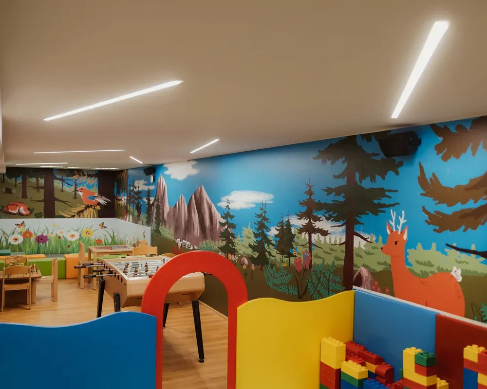 Colorful playroom with a vibrant forest-themed mural featuring trees and animals. Play tables, a foosball table, and building blocks create a playful atmosphere.