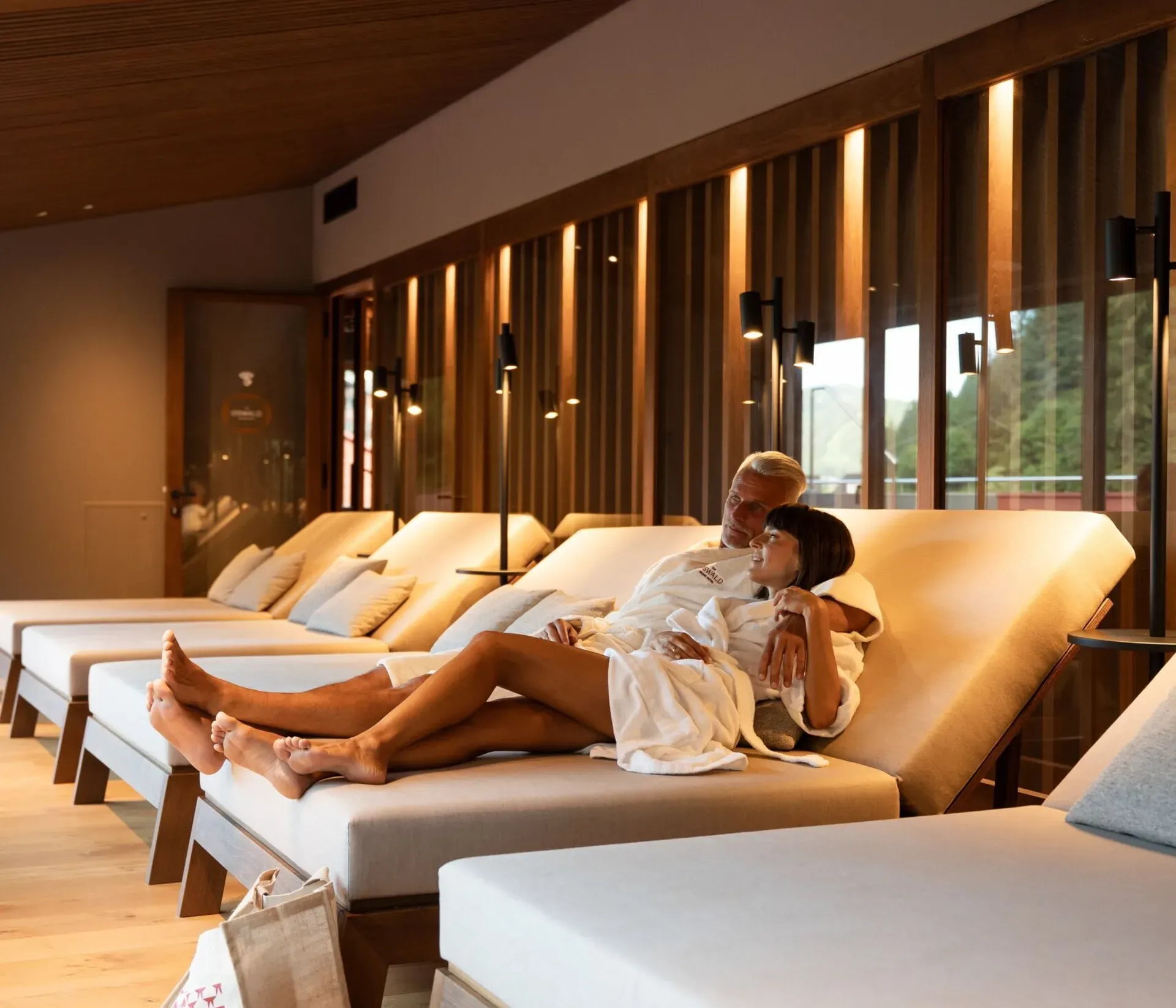 Ein eleganter Ruhebereich im The Oswald Grand Hotel in Wolkenstein in Gröden mit bequemen, ergonomisch geformten Liegen und warmem, indirektem Licht. Ein Paar entspannt gemeinsam in Bademänteln auf einer der Liegen. Die Einrichtung aus Holz, die gedeckten Naturtöne und die sanfte Beleuchtung schaffen eine ruhige und behagliche Atmosphäre im Wellnessbereich.