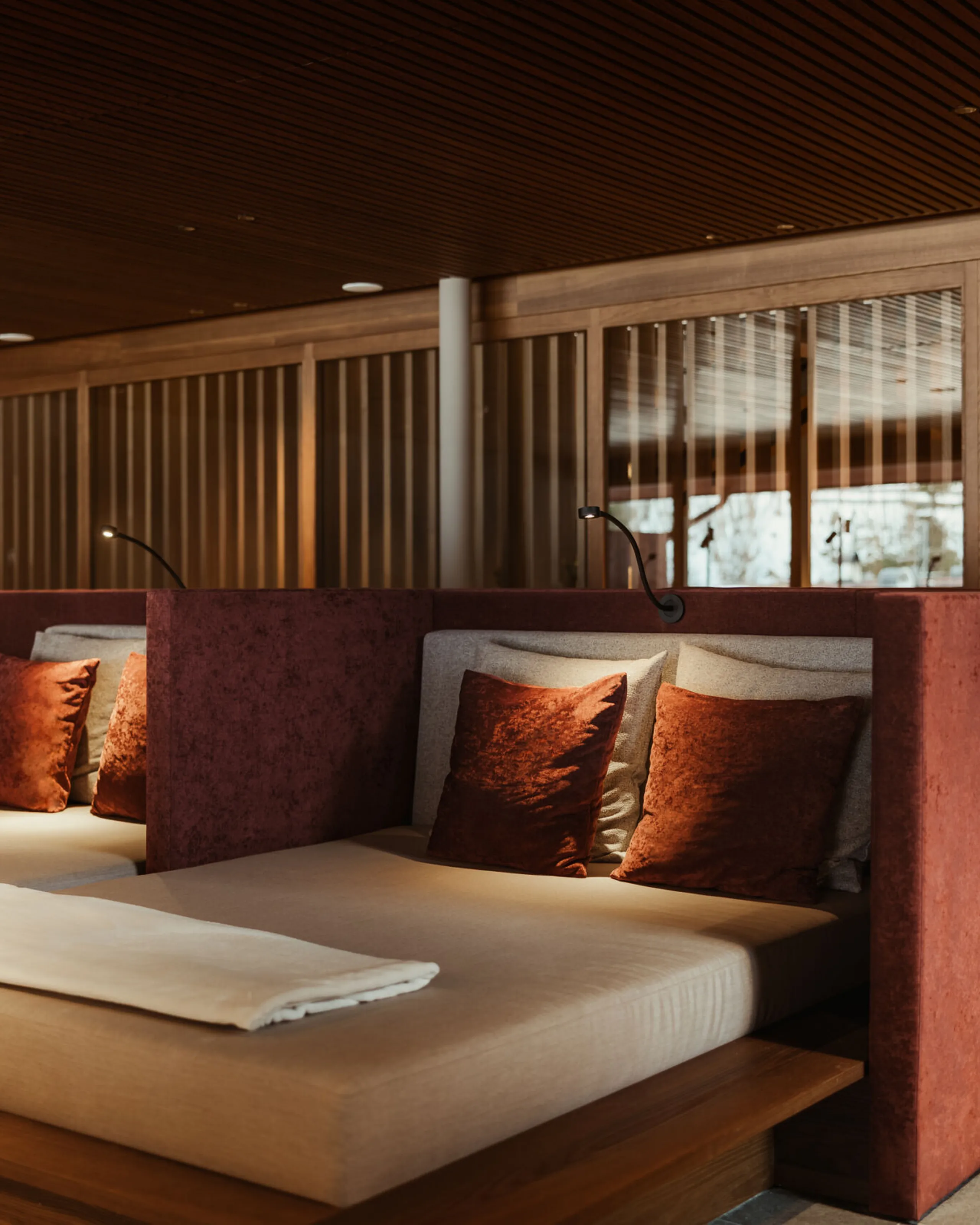 Luxurious spa room with plush daybeds featuring rust-colored cushions, soft lighting, and wooden partitions. A potted plant adds greenery for relaxation.